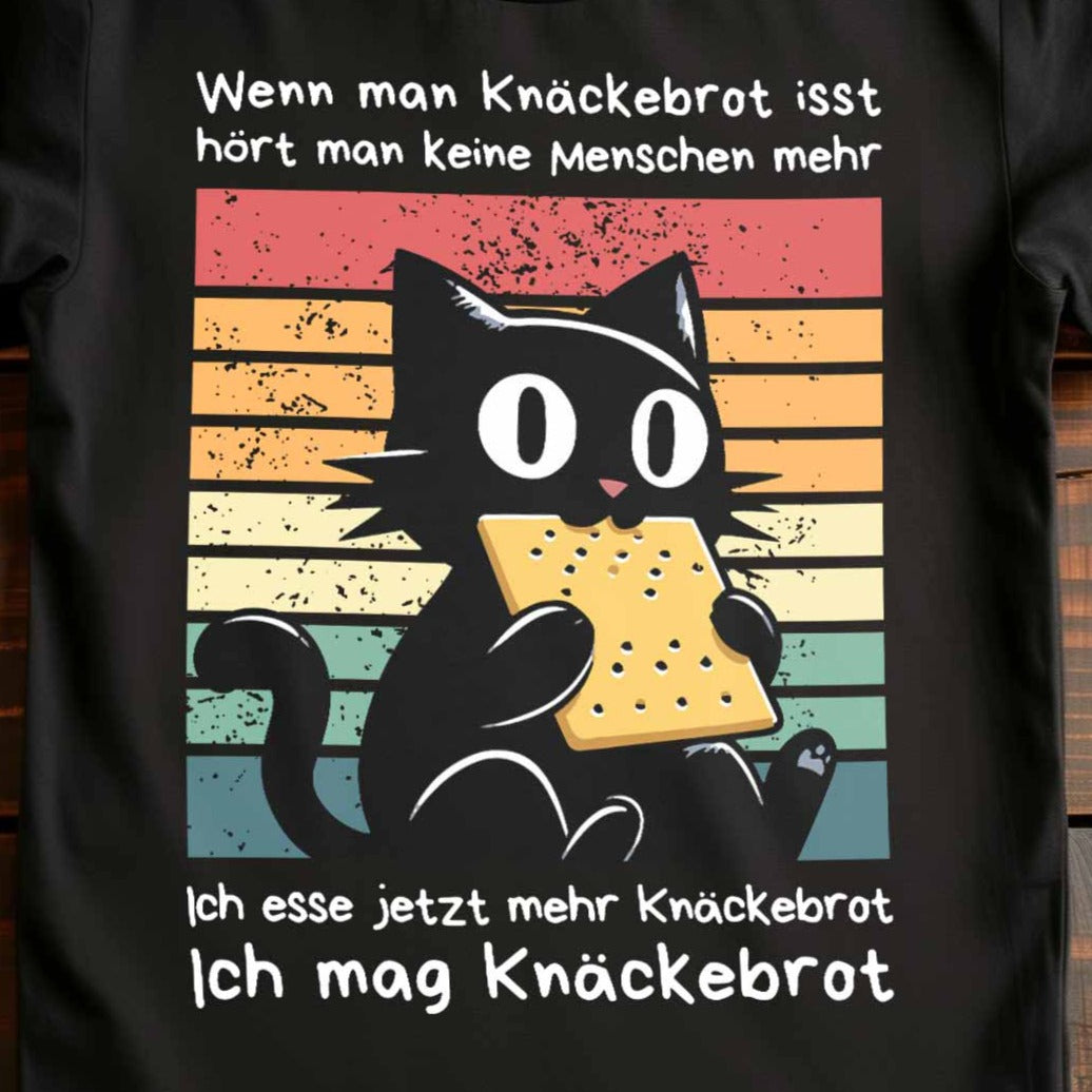 Nahaufnahme eines schwarzen Damen-Shirts auf Holzboden mit Design einer schwarzen Katze und dem Spruch 'Ich mag Knäckebrot'