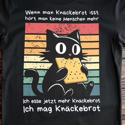 Nahaufnahme eines schwarzen T-Shirts auf Holzboden mit Design einer schwarzen Katze und dem Spruch 'Ich mag Knäckebrot'