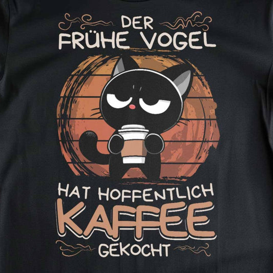 Nahaufnahme eines schwarzen UNISEX T-Shirts auf Holzboden mit Design einer schwarzen Katze und dem lustigen Spruch 'Der frühe Vogel hat hoffentlich Kaffee gekocht'