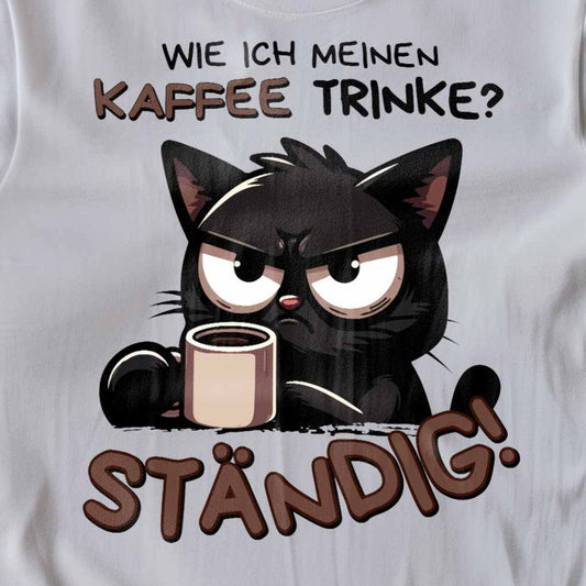 Nahaufnahme eines weißen T-Shirts mit Design einer Kaffee trinkenden schwarzen Katze und lustigen Spruch: 'Wie ich meinen Kaffee trinke? Ständig!'