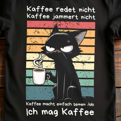 Nahaufnahme eines schwarzen Damen-Shirts auf Holzboden mit Kaffee trinkender Katze und dem lustigen Spruch 'Ich mag Kaffee'