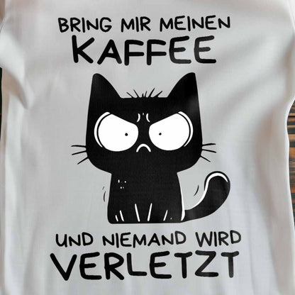 Nahaufnahme eines weißen Damen-Shirts auf Holzboden mit Katze und Spruch 'Bring mir meinen Kaffee und niemand wird verletzt'