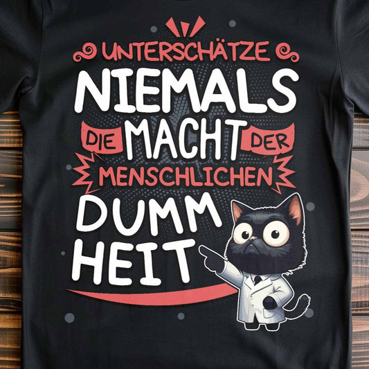 Nahaufnahme eines schwarzen T-Shirts auf Holzboden mit Design einer Katze als Doktor mit dem lustigen Spruch 'Unterschätze niemals die Macht der menschlichen Dummheit'