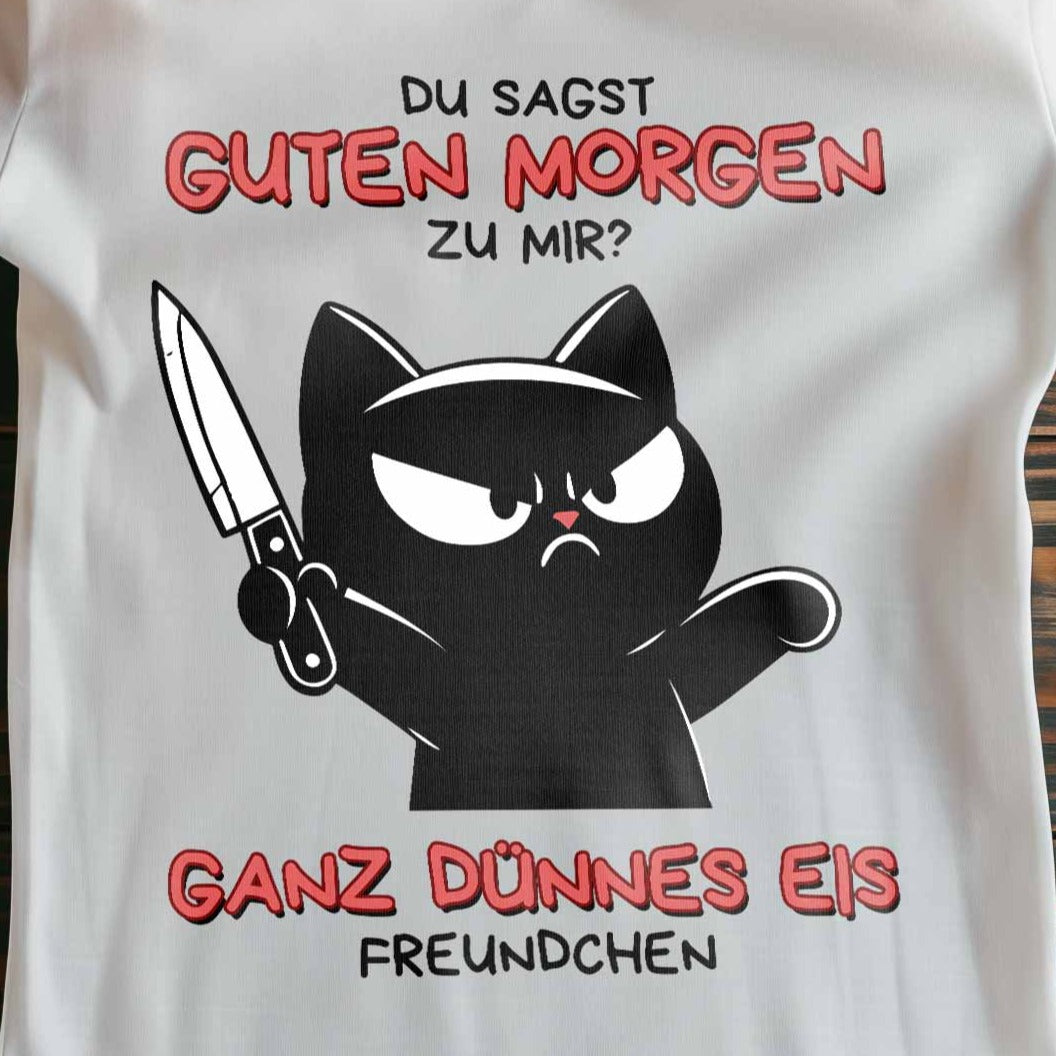 Nahaufnahme eines weißen Damen-T-Shirts auf Holzboden mit Design einer schwarzen Katze mit Messer und dem lustigen Spruch 'Guten Morgen? Ganz dünnes Eis'