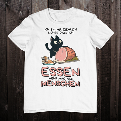 Weißes T-Shirt auf einem Holzboden zeigt ein Design einer schwarzen Katze mit einem Schweinebraten und dem lustigen Spruch: 'Ich bin mir ziemlich sicher, dass ich Essen mehr mag als Menschen'